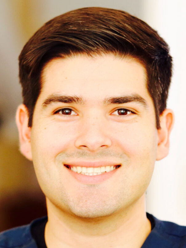 Close-up portrait of a smiling young man wearing navy scrubs.