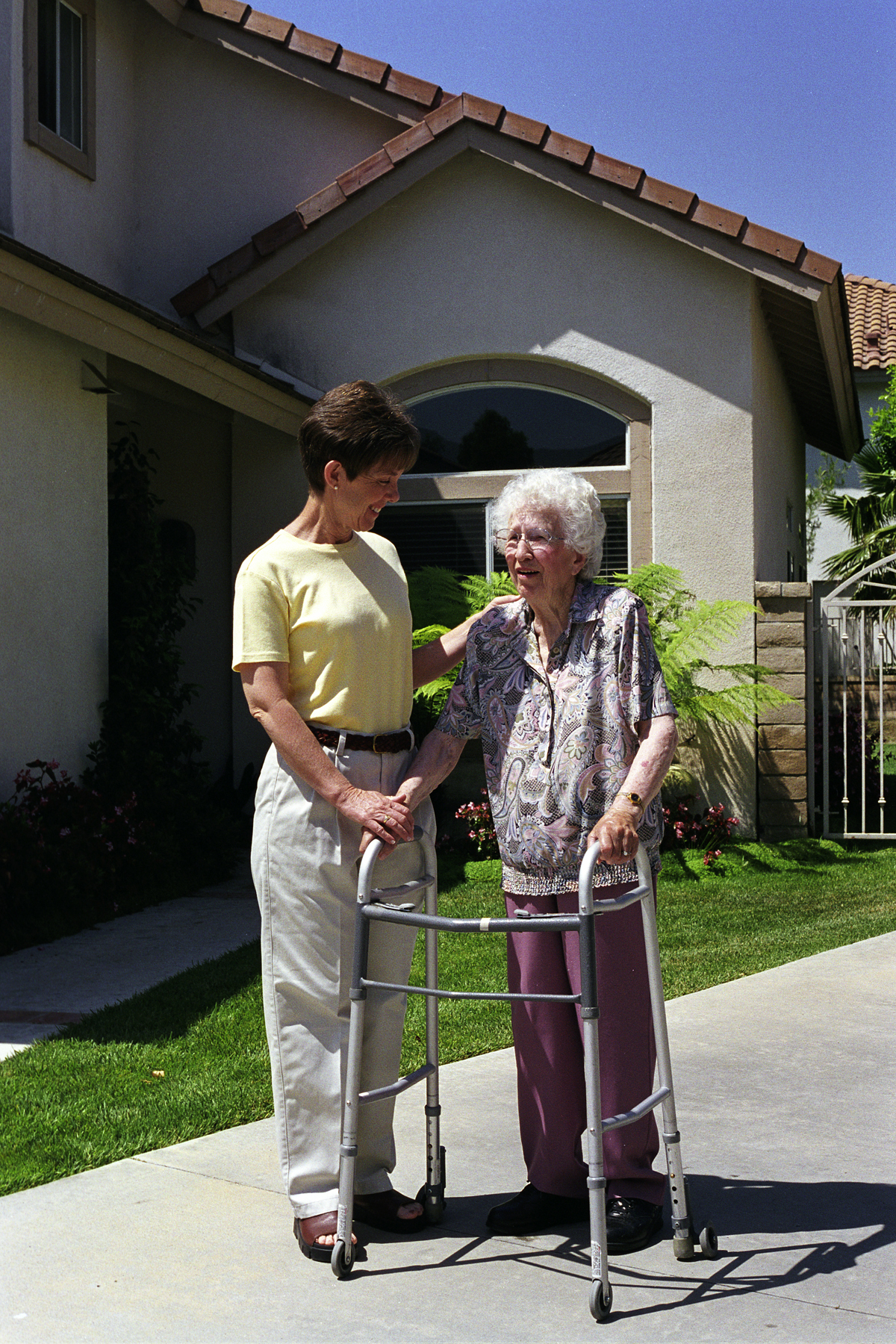 Caregiver in a yellow shirt assisting an elderly woman with a walker on a driveway outside a house.