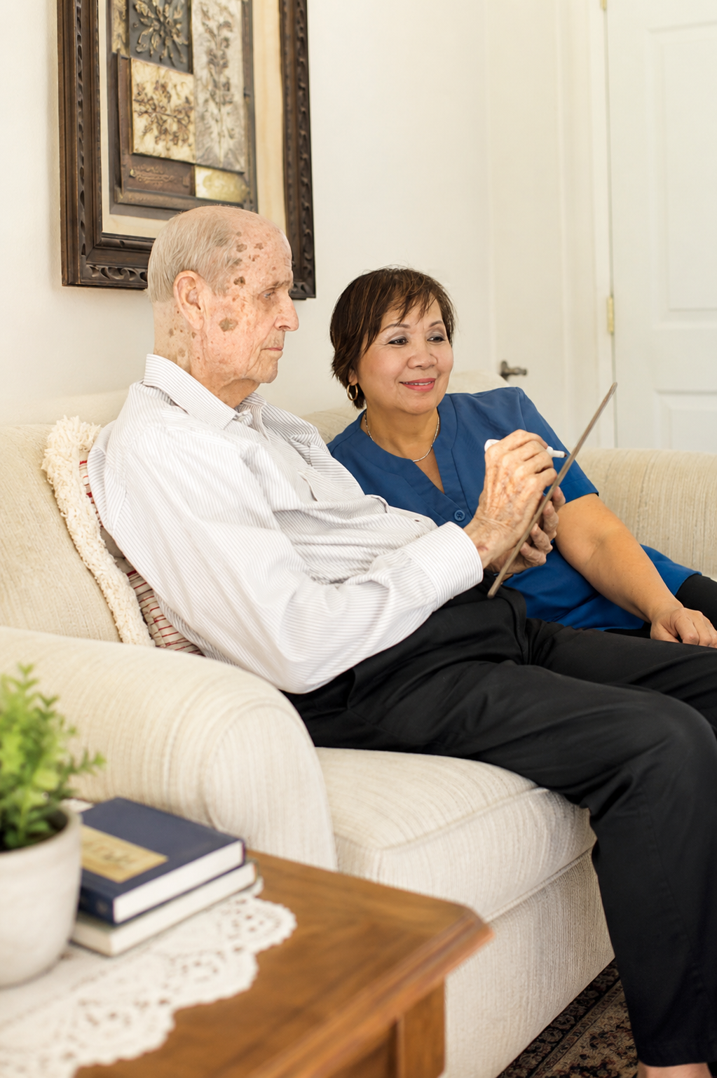 Caregiver in blue scrubs helping an elderly man use a tablet while sitting together on a couch at home.