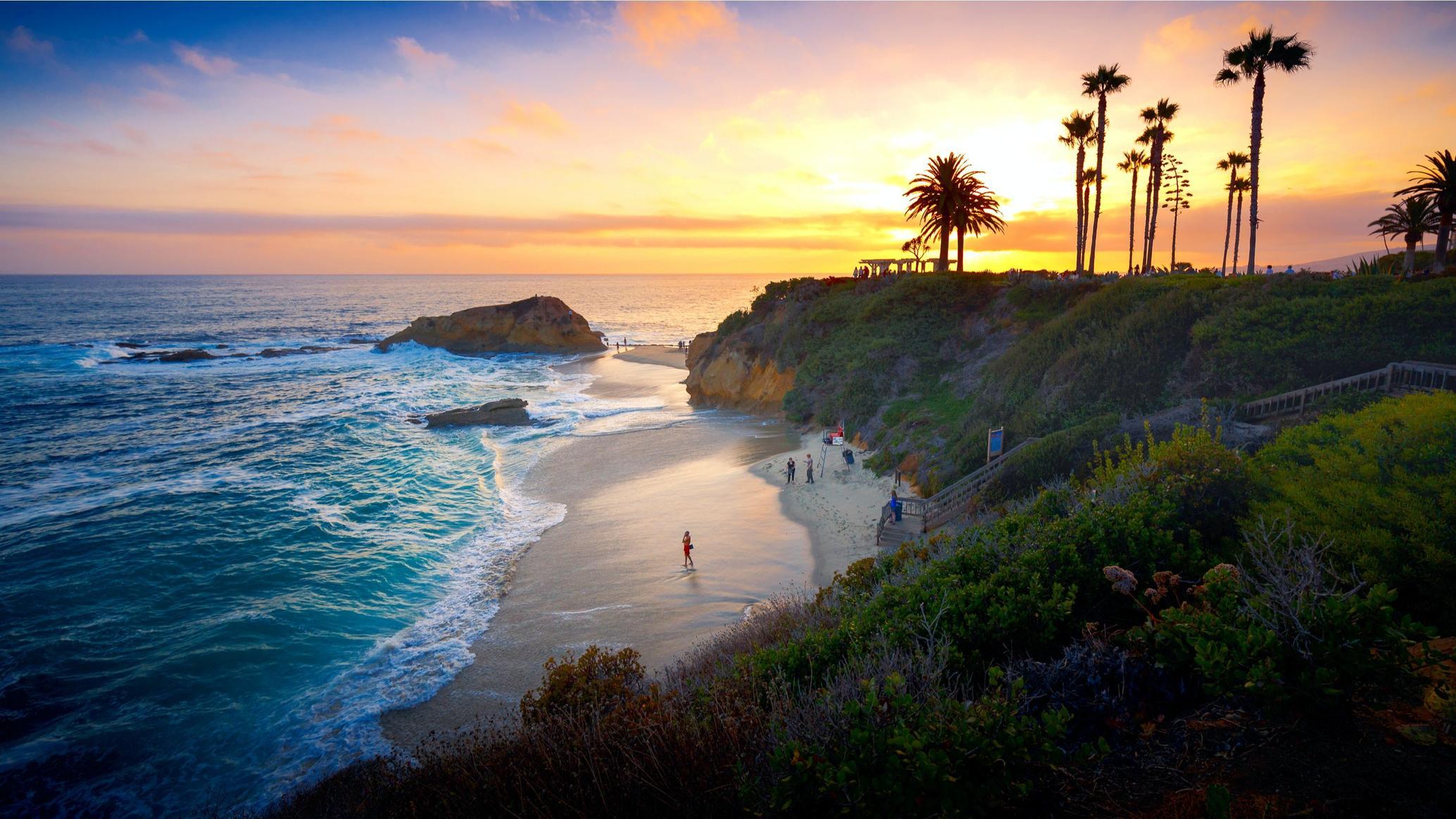 Scenic coastal cove at sunset with palm trees silhouetted on the cliffside above turquoise waves.
