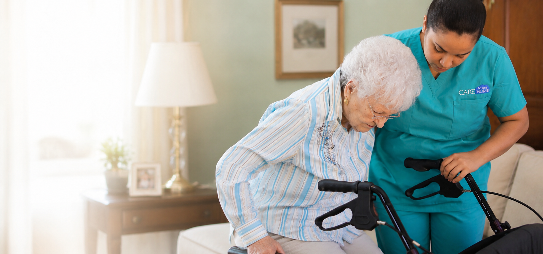 Caregiver in teal scrubs helping an elderly woman rise from a couch using a rollator walker in a living room.