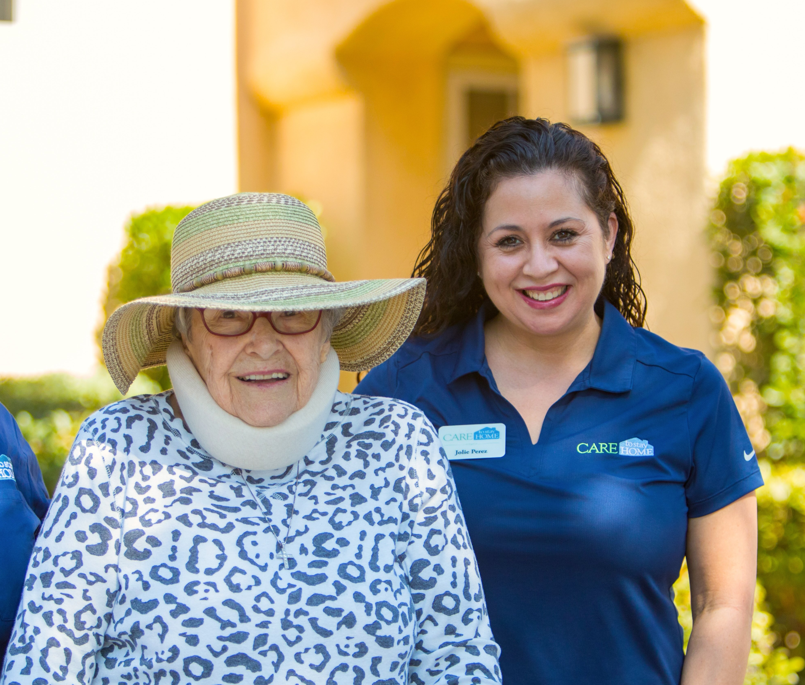 Smiling elderly woman in a sun hat standing outdoors beside a caregiver in a navy polo shirt.