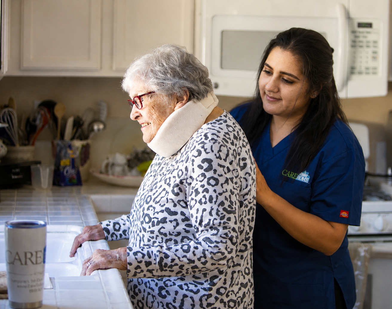 Caregiver in navy scrubs assisting an elderly woman wearing a neck brace at a kitchen counter.