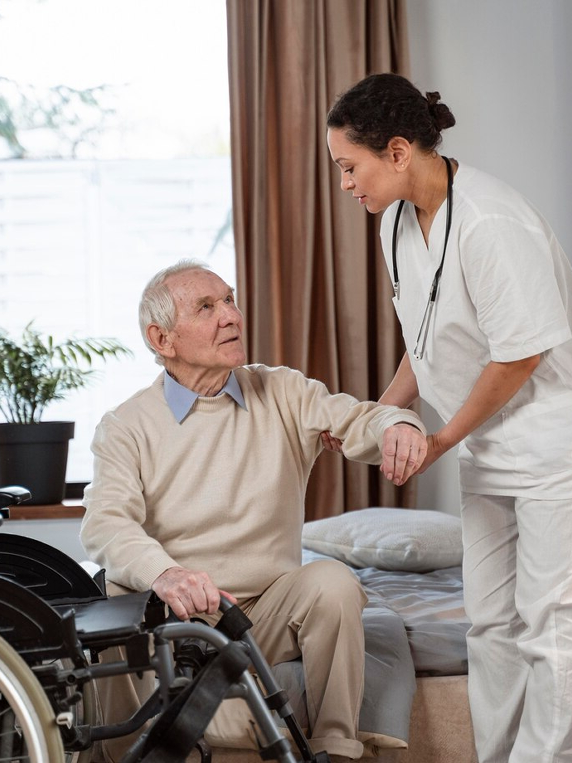 Nurse in white uniform with stethoscope helping an elderly man transfer from a bed to a wheelchair.