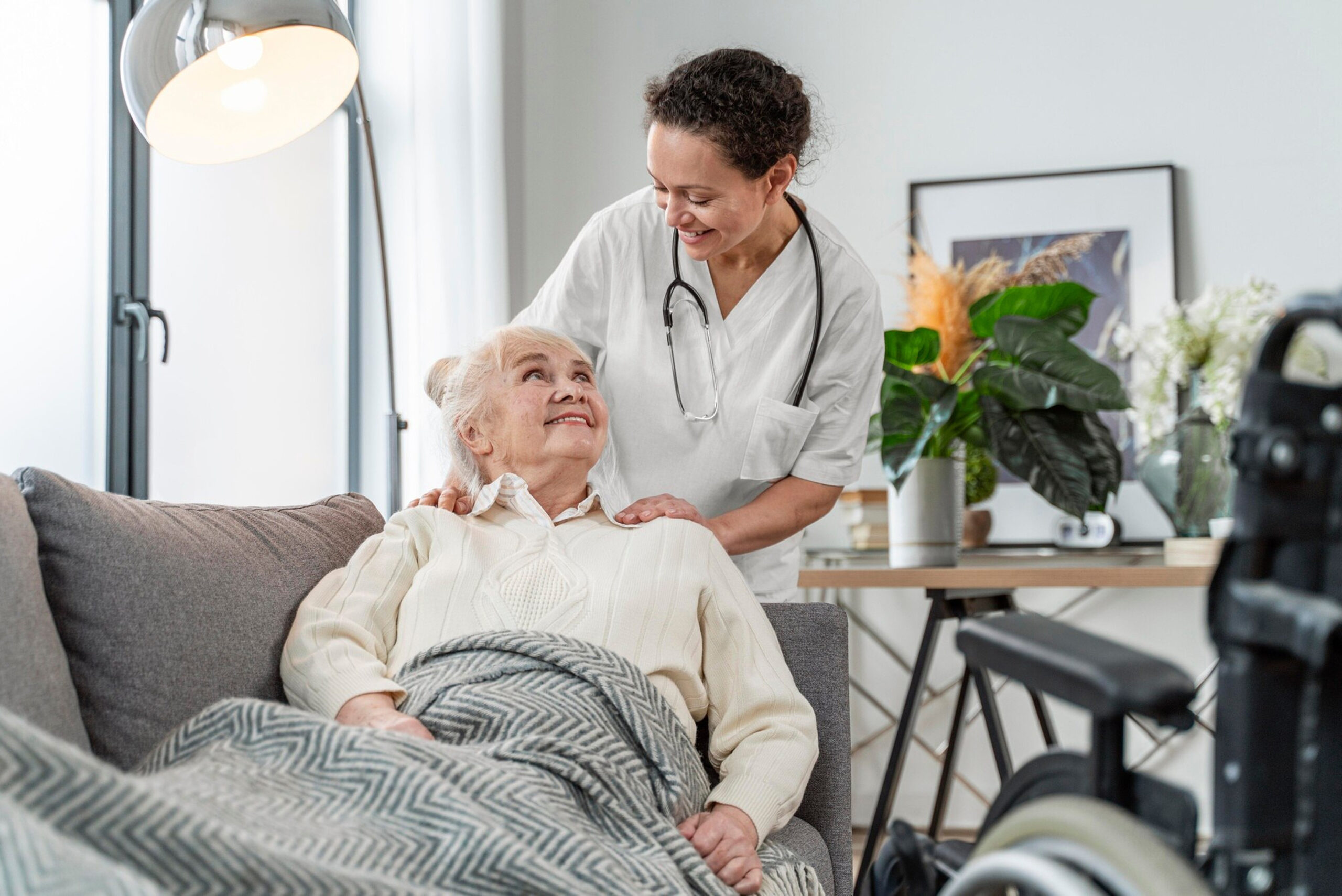 Nurse in white uniform with stethoscope comforting an elderly woman resting on a sofa, wheelchair visible nearby.