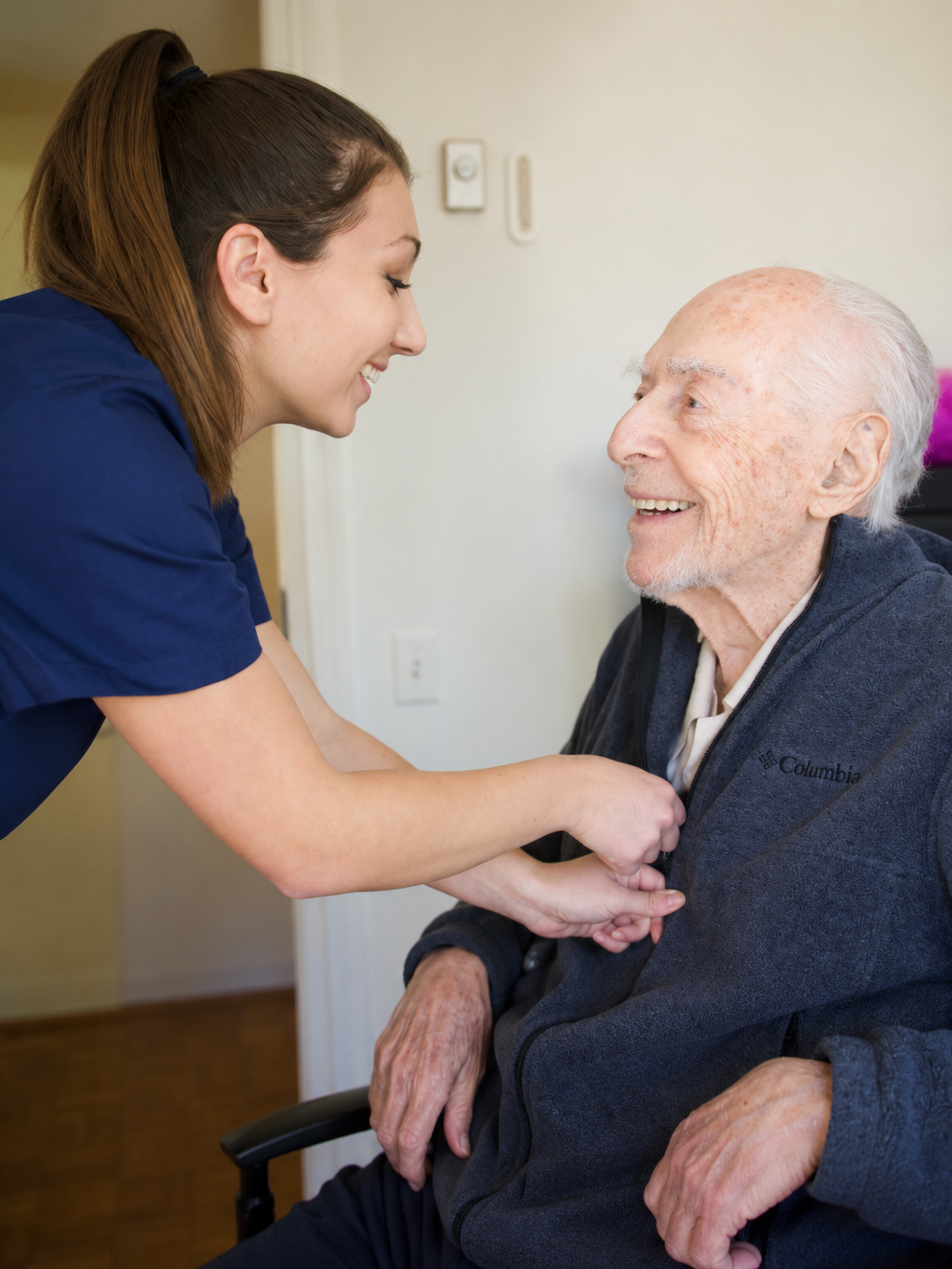 Caregiver in navy scrubs smiling and holding hands with an elderly man seated in a wheelchair.