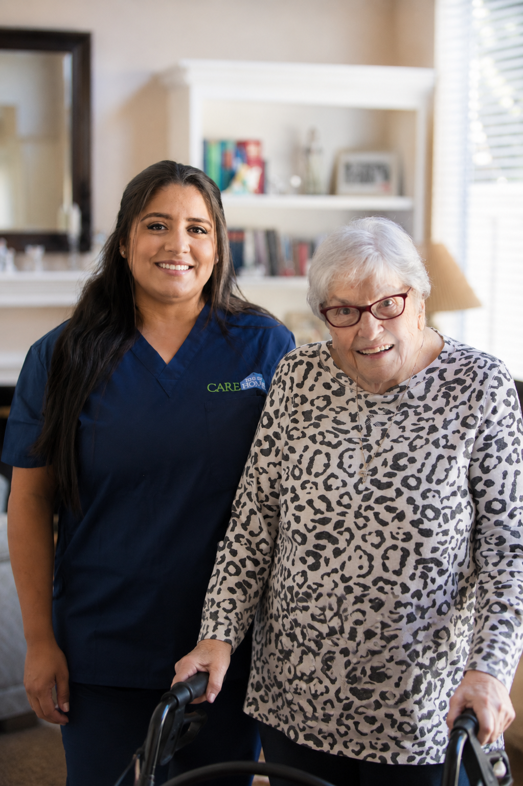 Smiling caregiver in navy scrubs standing beside an elderly woman using a walker at home.