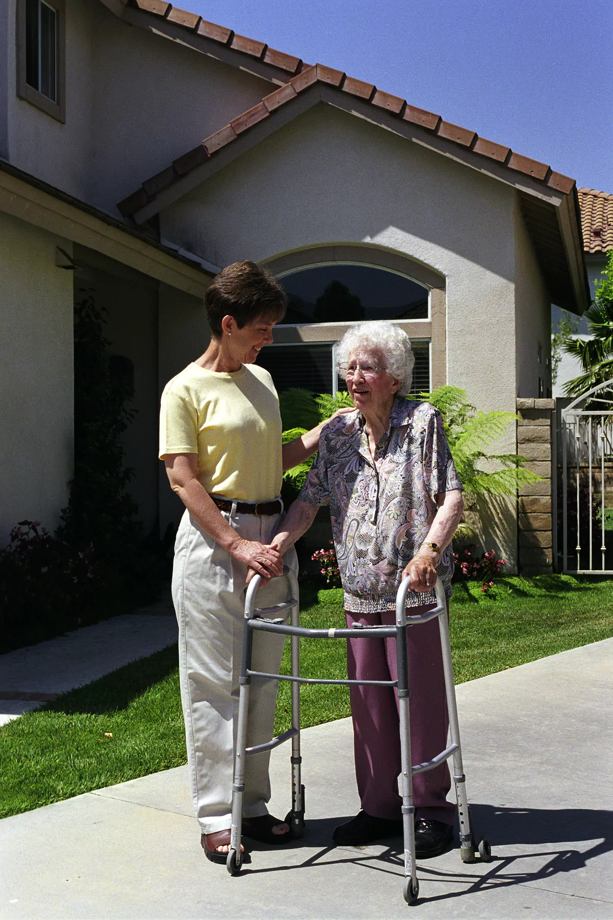 Caregiver in a yellow shirt assisting an elderly woman with a walker on a driveway outside a house.
