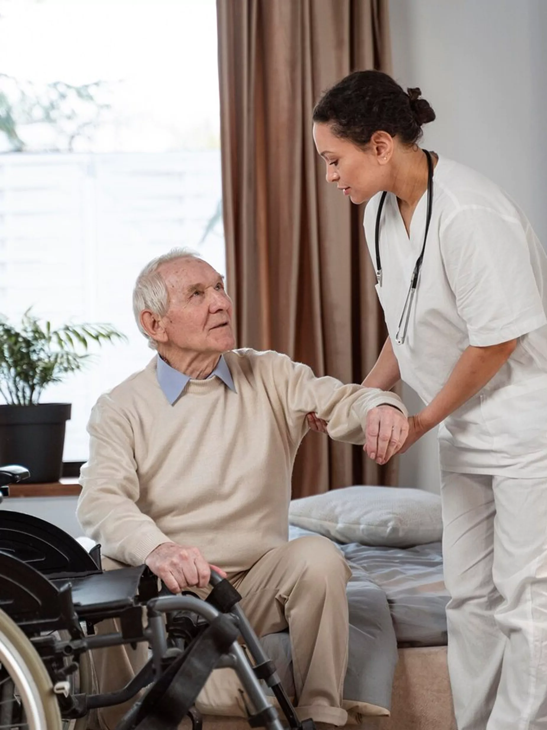 Nurse in white uniform with stethoscope helping an elderly man transfer from a bed to a wheelchair.