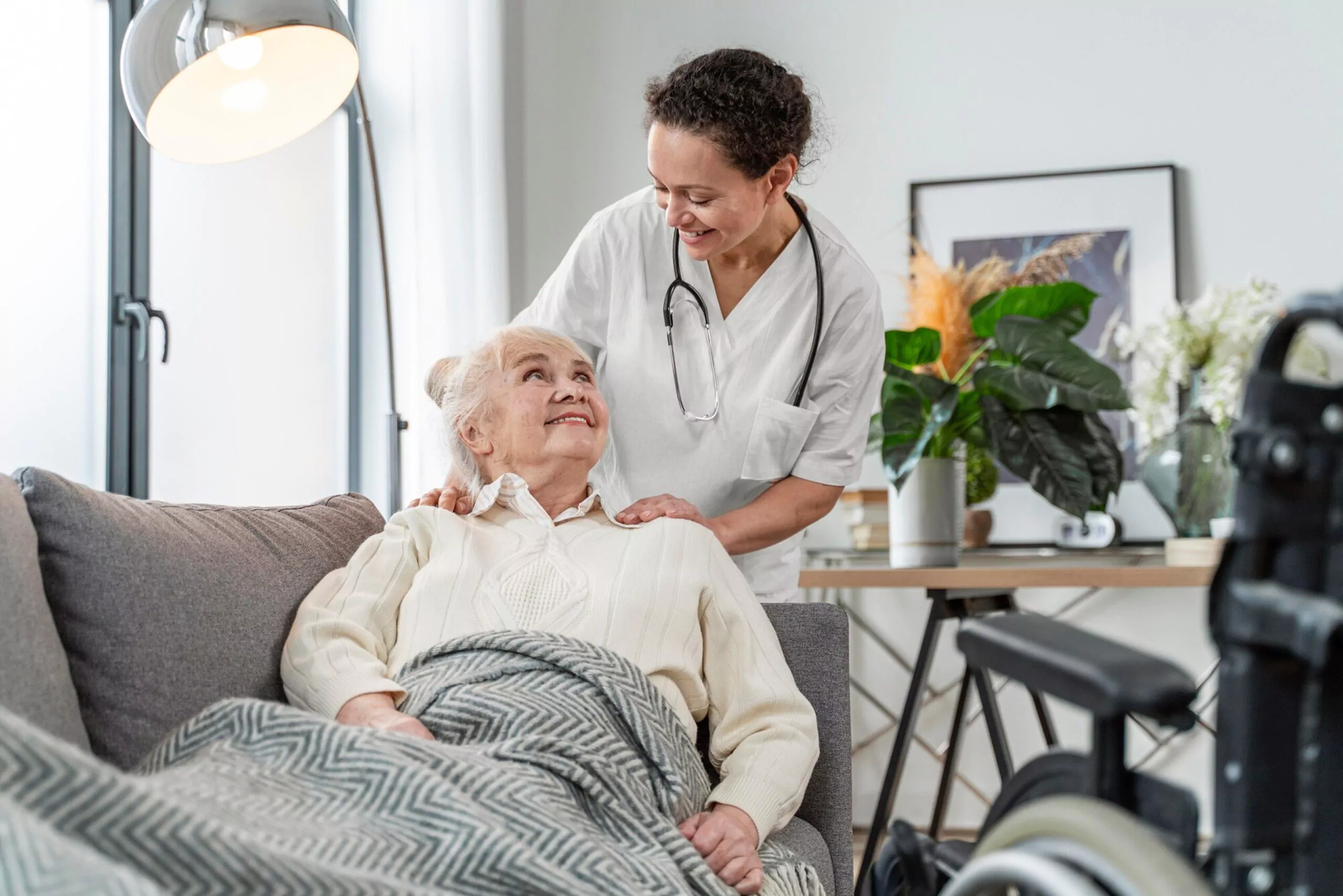 Nurse in white uniform with stethoscope comforting an elderly woman resting on a sofa, wheelchair visible nearby.