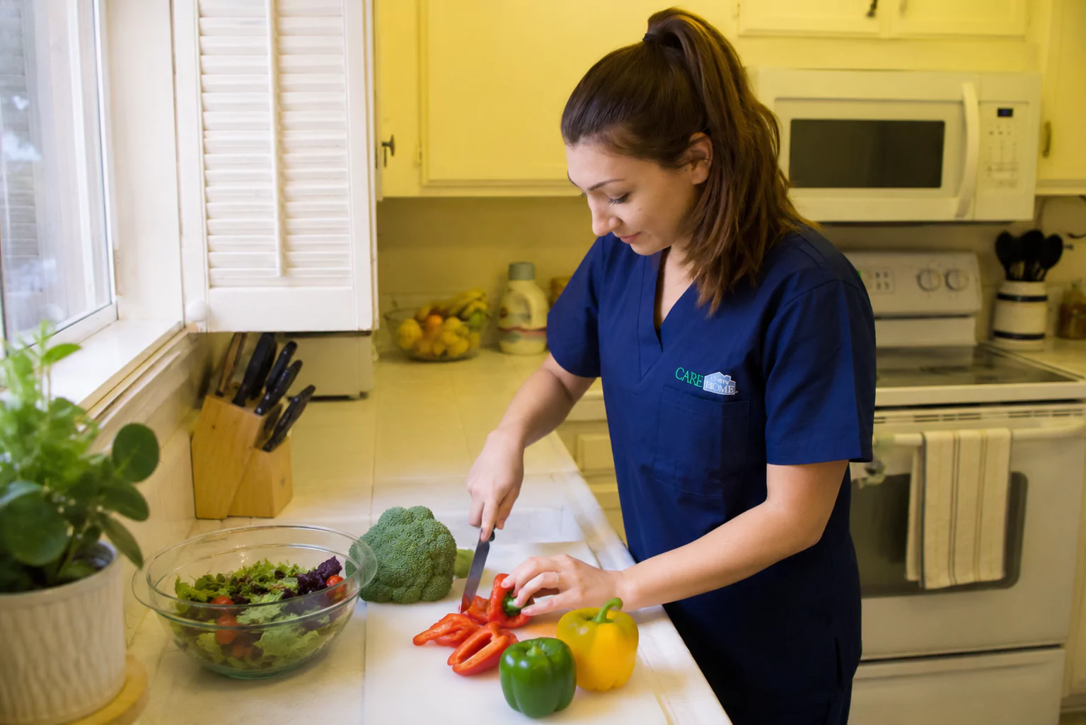 Caregiver in navy scrubs chopping fresh vegetables to prepare a meal in a home kitchen.