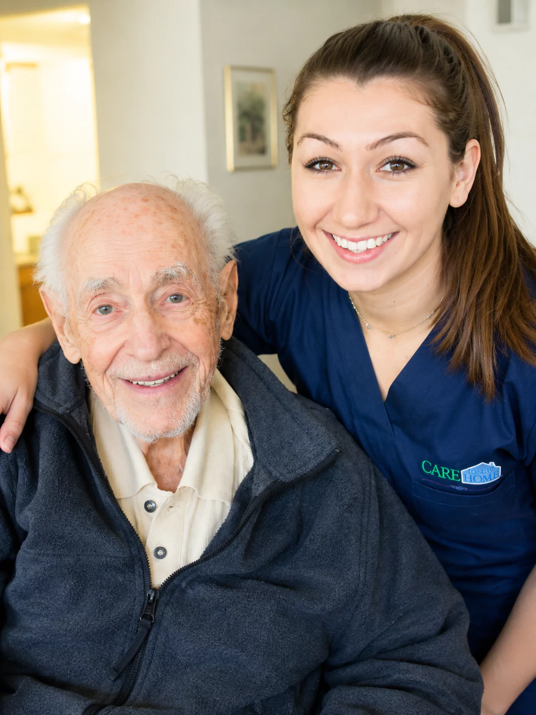 Smiling caregiver in navy scrubs posing beside an elderly man, both looking at the camera indoors.