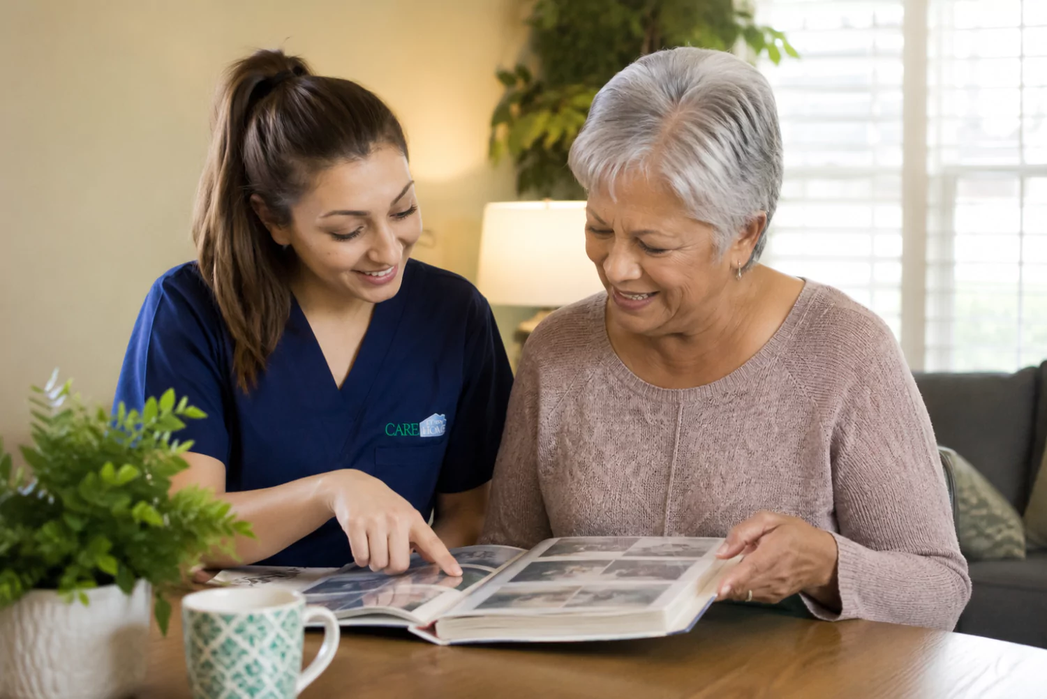 Caregiver in navy scrubs looking through a photo album together with a smiling elderly woman at home.